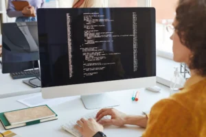Person typing on a keyboard with a large monitor showing lines of code in a bright office environment, seen from the side.